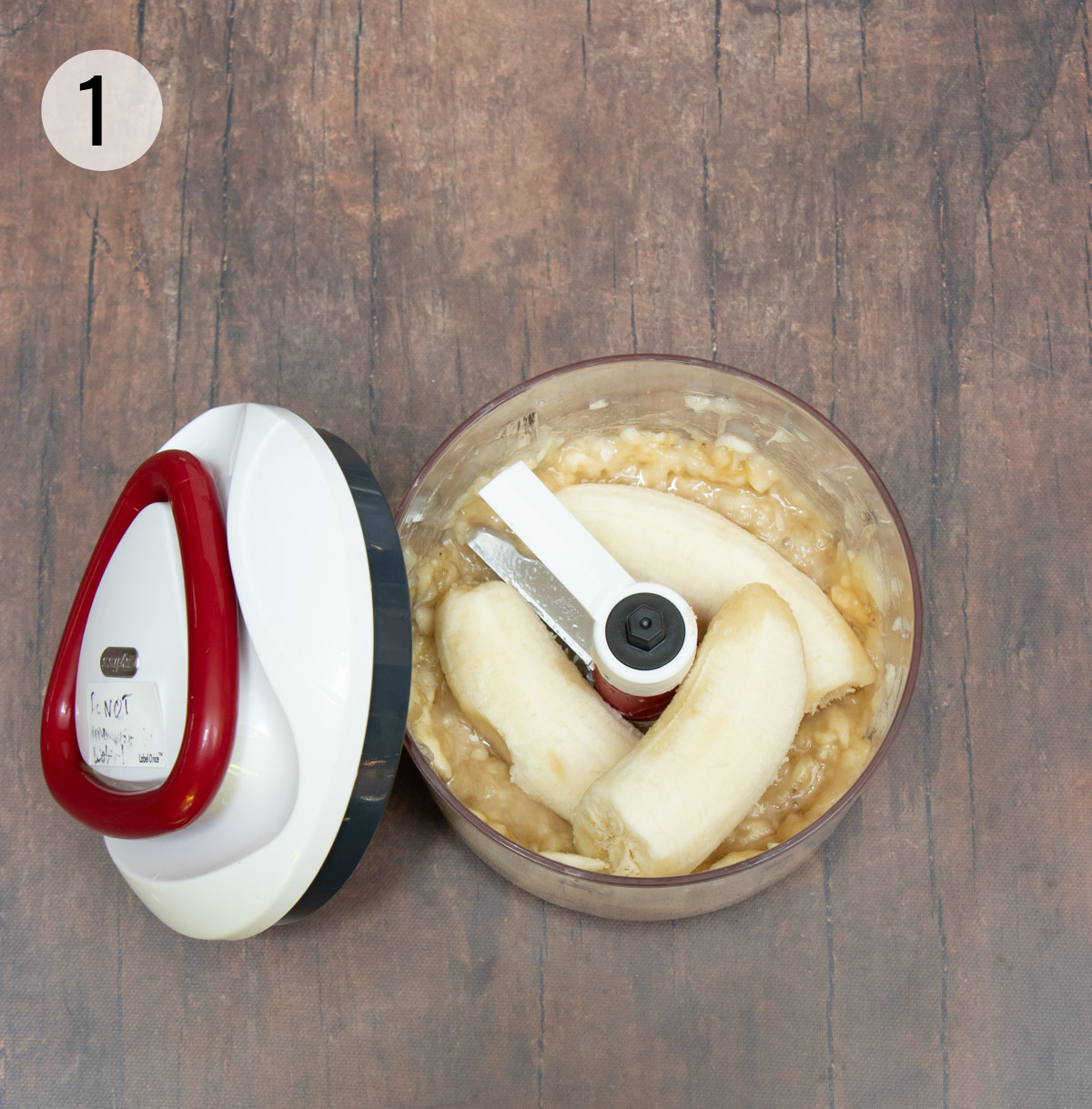 Handheld food chopper with bananas to puree and white lid with red handle on left side. 