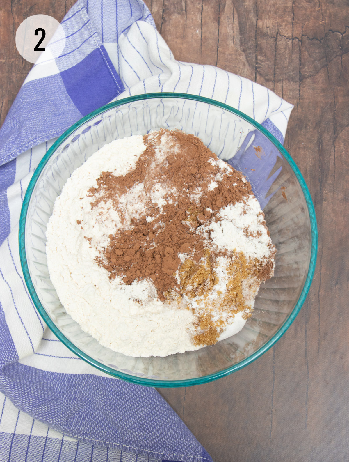 Glass bowl with flour, cocoa powder and brown sugar with blue and white towel draped on left side. 