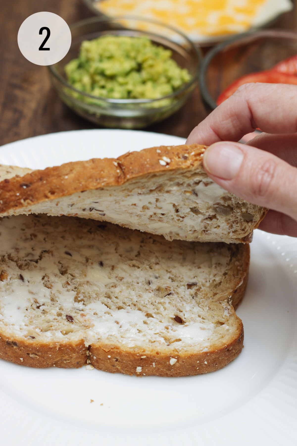 Hand lifting two buttered slices of bread with avocado in bowl in background.