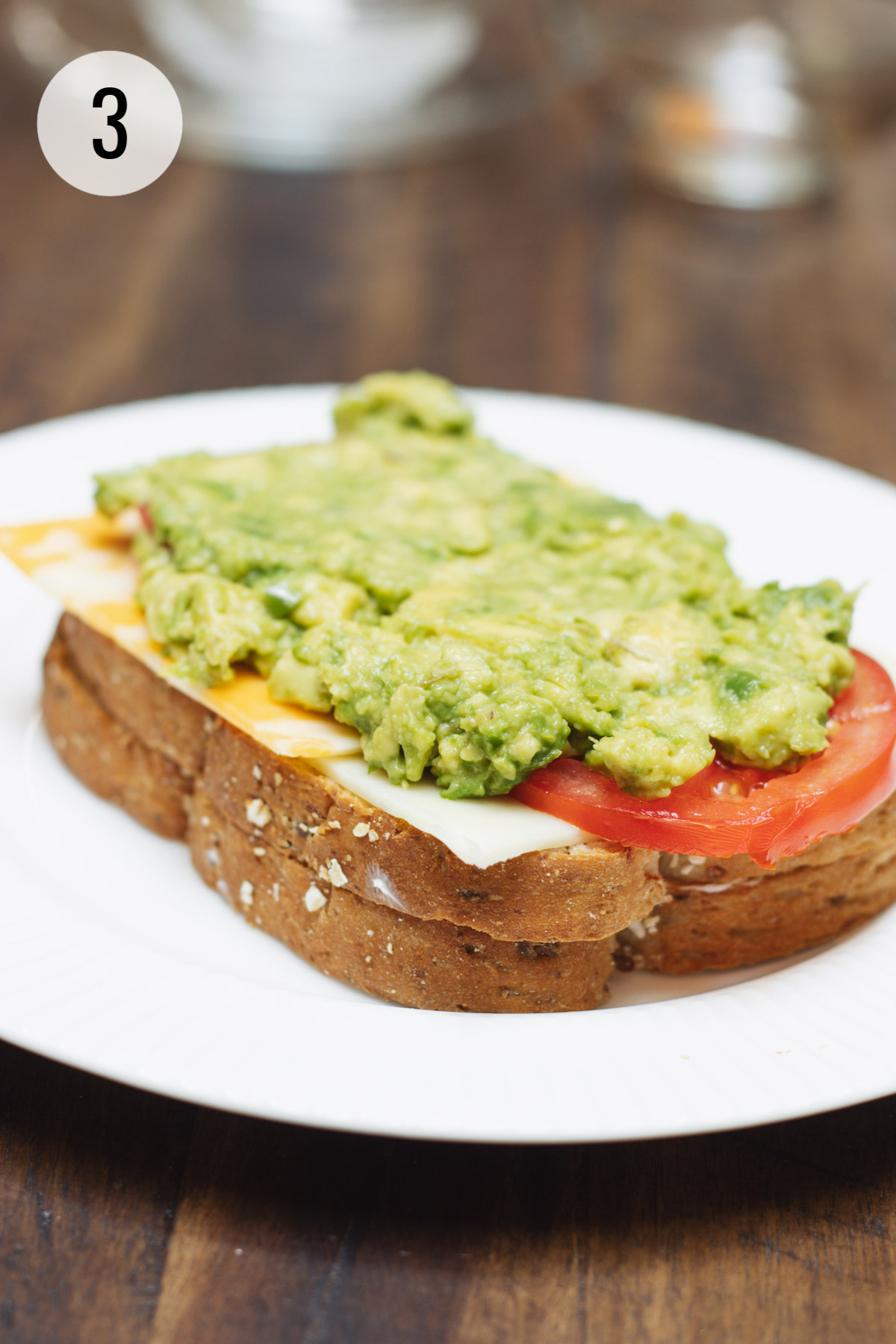 Two slices of bread topped with cheese, tomato and smashed avocado placed on a white plate.