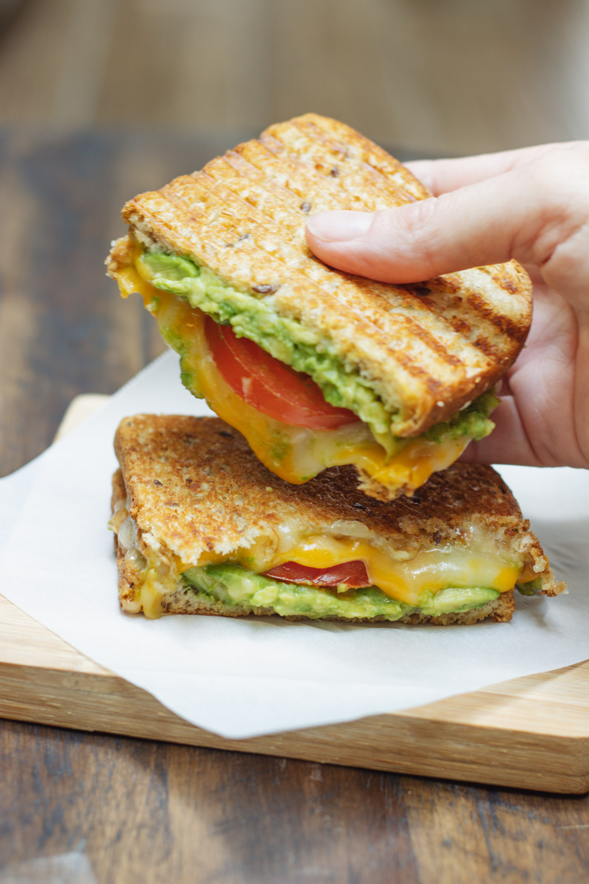 Two halves of a tomato and avocado grilled cheese sandwich on a wooden board with hand holding one half. 