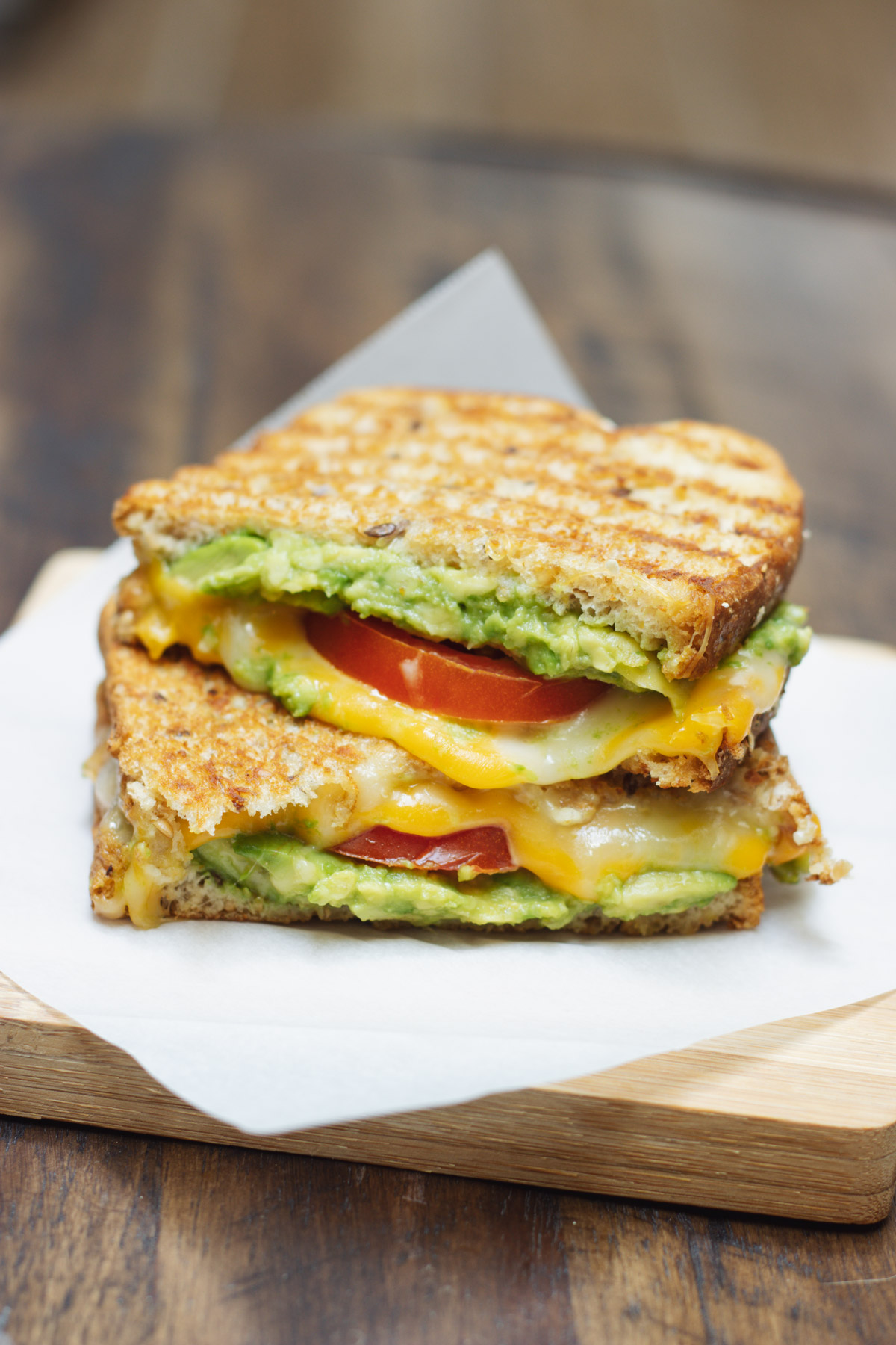 Two halves of a tomato and avocado grilled cheese sandwich stacked on parchment paper on a wooden board.