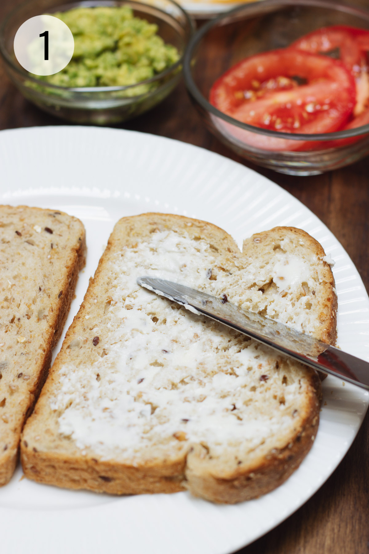 Knife spreading butter on a piece of whole grain bread with tomato and avocado in background. 