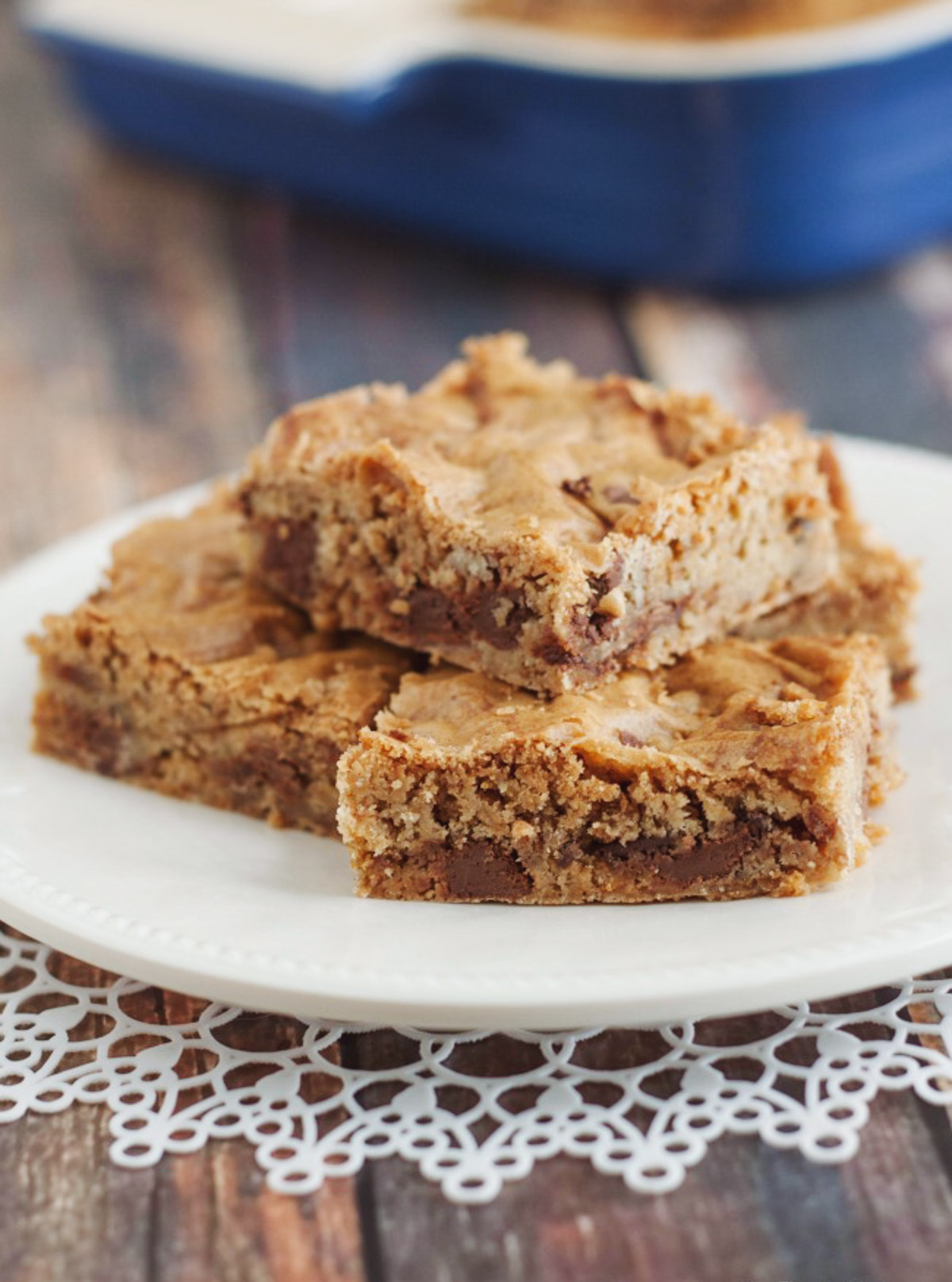 Stack of blondie bars with chocolate chips on a white plate on white doily with blue baking pan in background. 