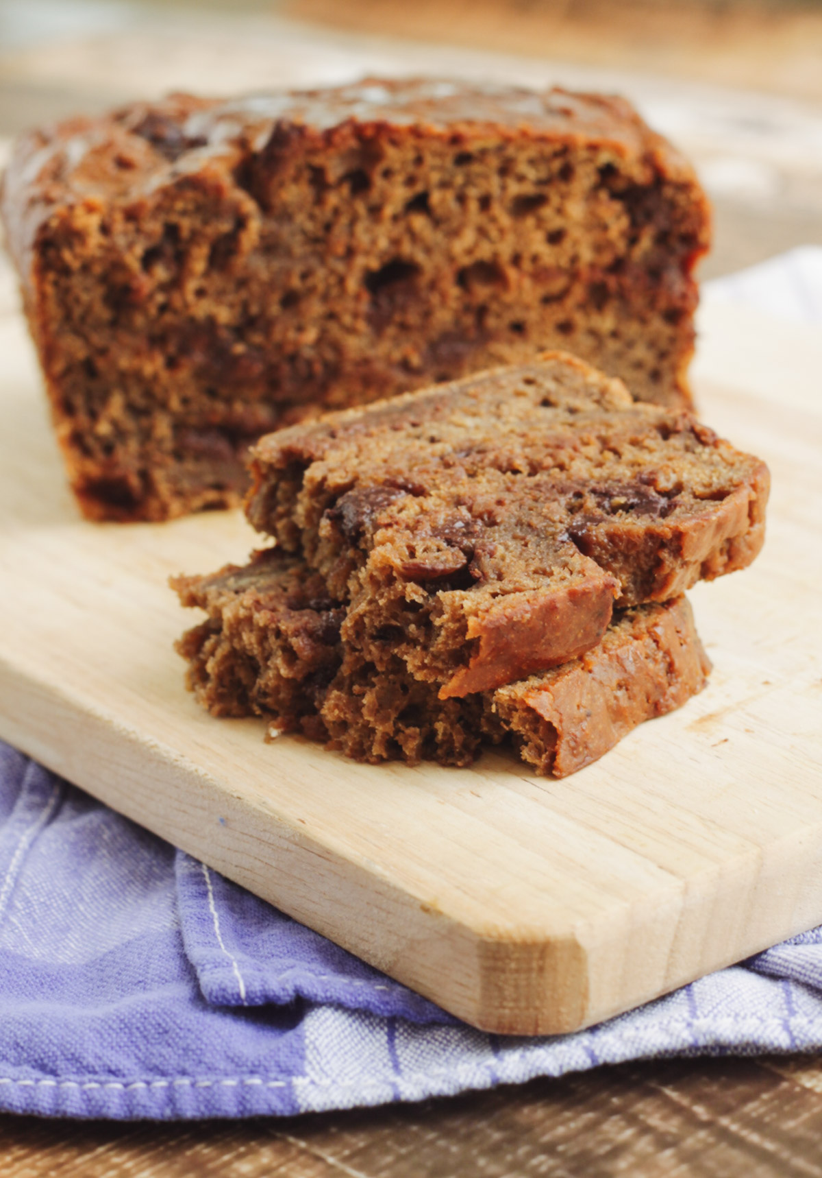 Two slices of chocolate chip banana bread with full loaf in background on a wooden cutting board and blue towel. 