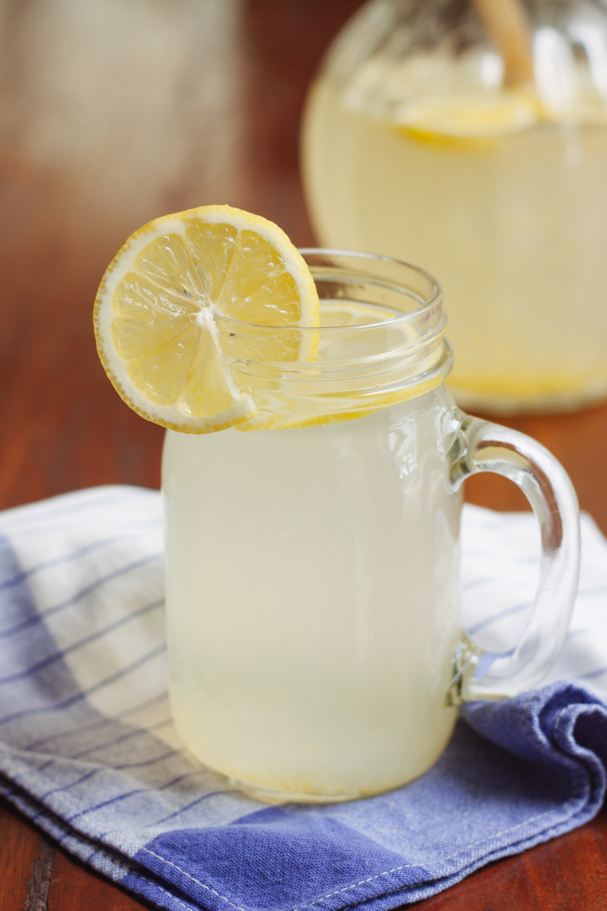 Glass mug with lemonade and lemon on top on a blue and white towel with pitcher of lemonade in background.