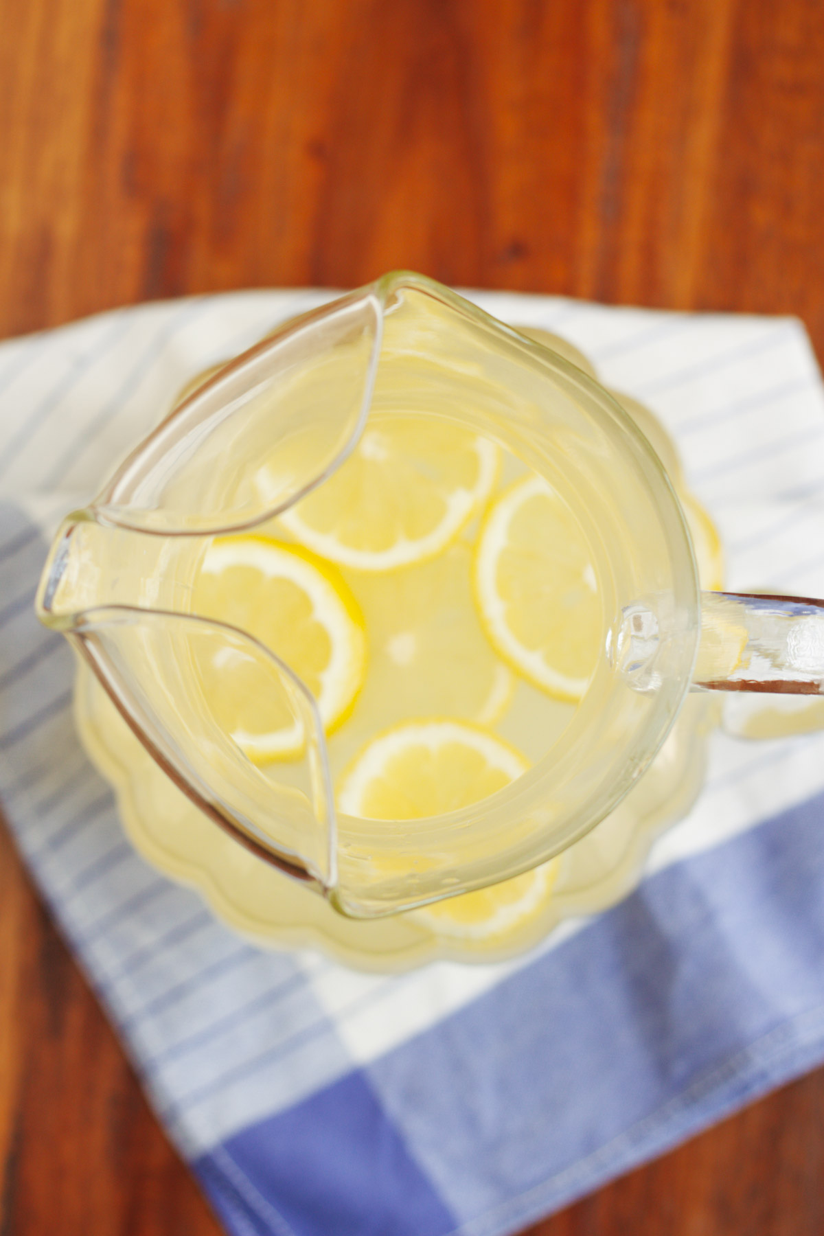 Top view image of a pitcher of lemonade with sliced lemons floating at top on a blue and white towel. 