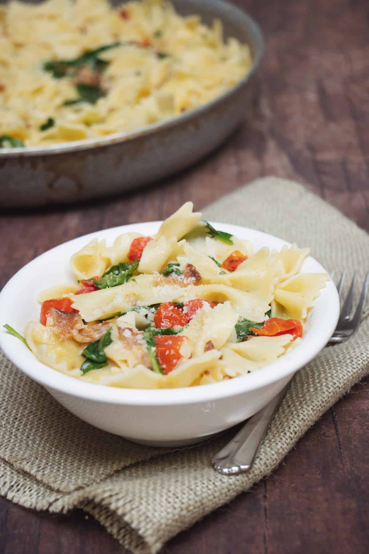 Bacon Spinach and Tomato pasta in a white bowl on brown napkin with extra in skillet in upper left corner.