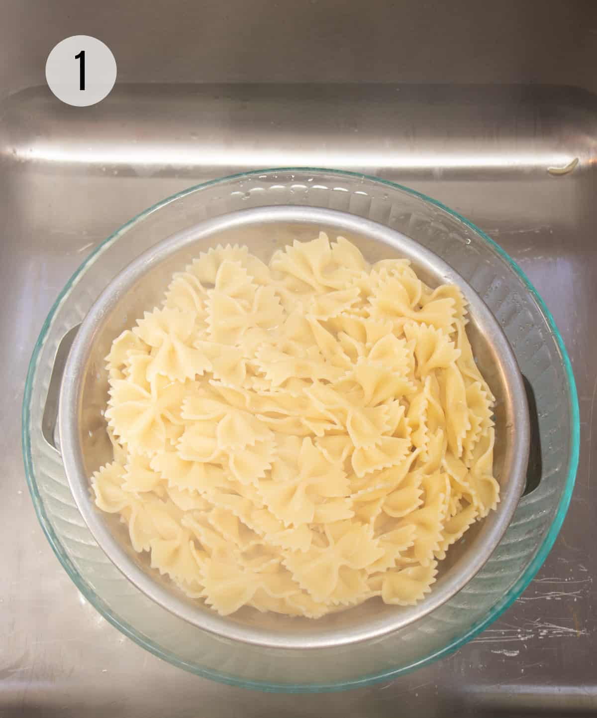 Colander of farfalle pasta draining in a clear bowl set in stainless steel sink. 