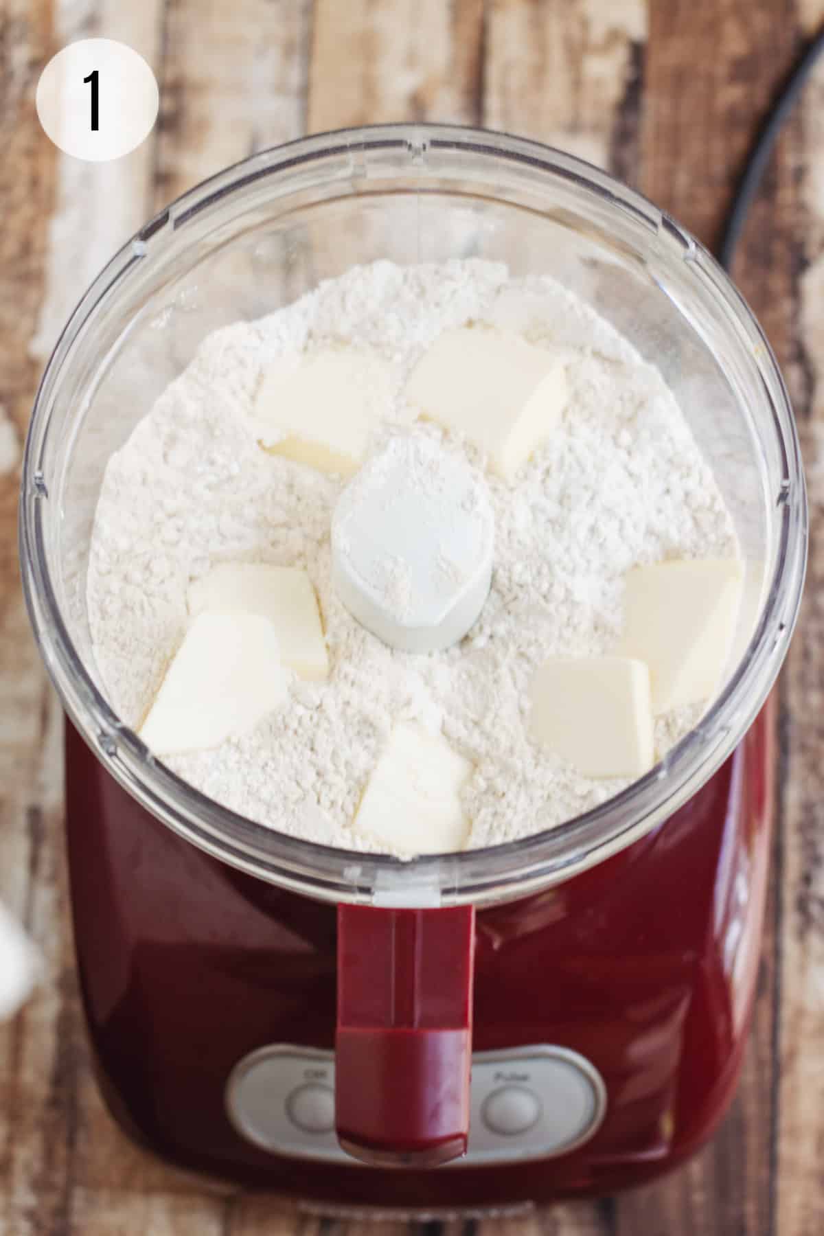 Food processor bowl with whole wheat biscuit mixture and squares of butter on a red processor base.