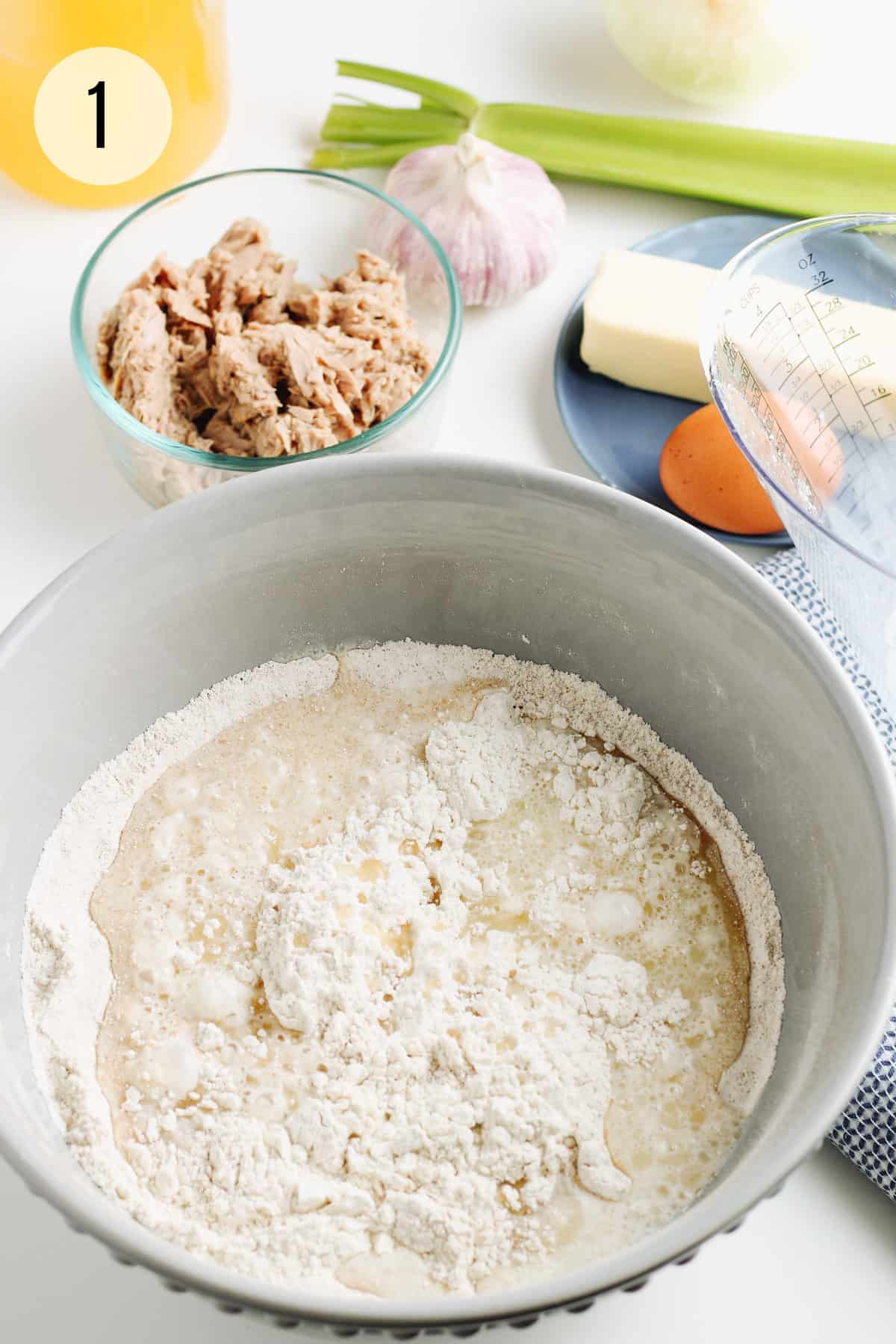 Grey bowl with baking powder biscuit dough ingredients plus tuna, garlic, celery stalk, butter and egg in upper background. 