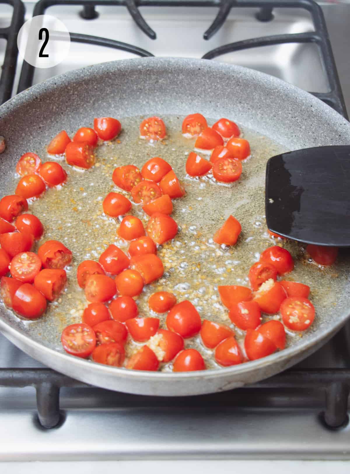 Sliced grape tomatoes sauteing in bacon fat in a grey skillet with black spatula. 