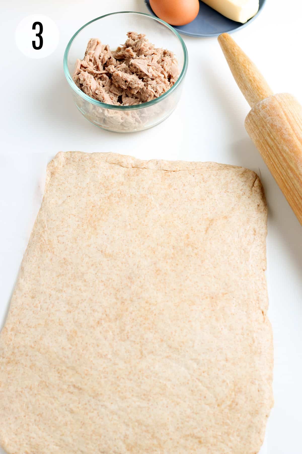 Rolled out whole wheat biscuit dough with wooden rolling pin, bowl of tuna and egg in upper and right background. 