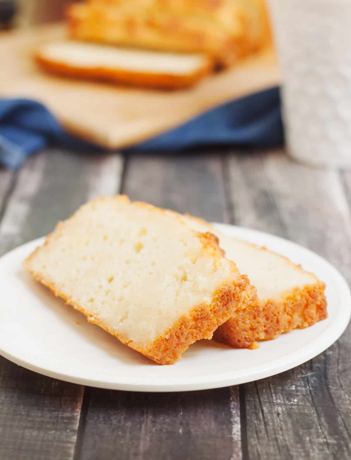 Two slices of lemon bread on a white plate with loaf of bread in background on blue napkin. 