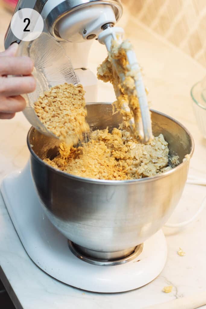 Pouring chopped peanuts into dough for Cracker Jack Rice Krispie Cookies in silver bowl of stand mixer. 