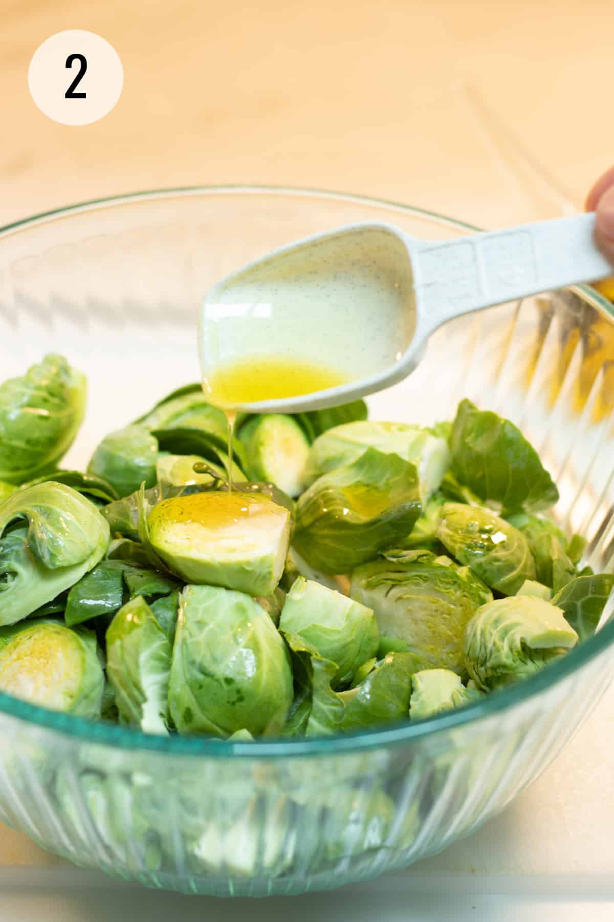 White measuring spoon drizzling olive oil in glass bowl full of cut Brussels sprouts. 