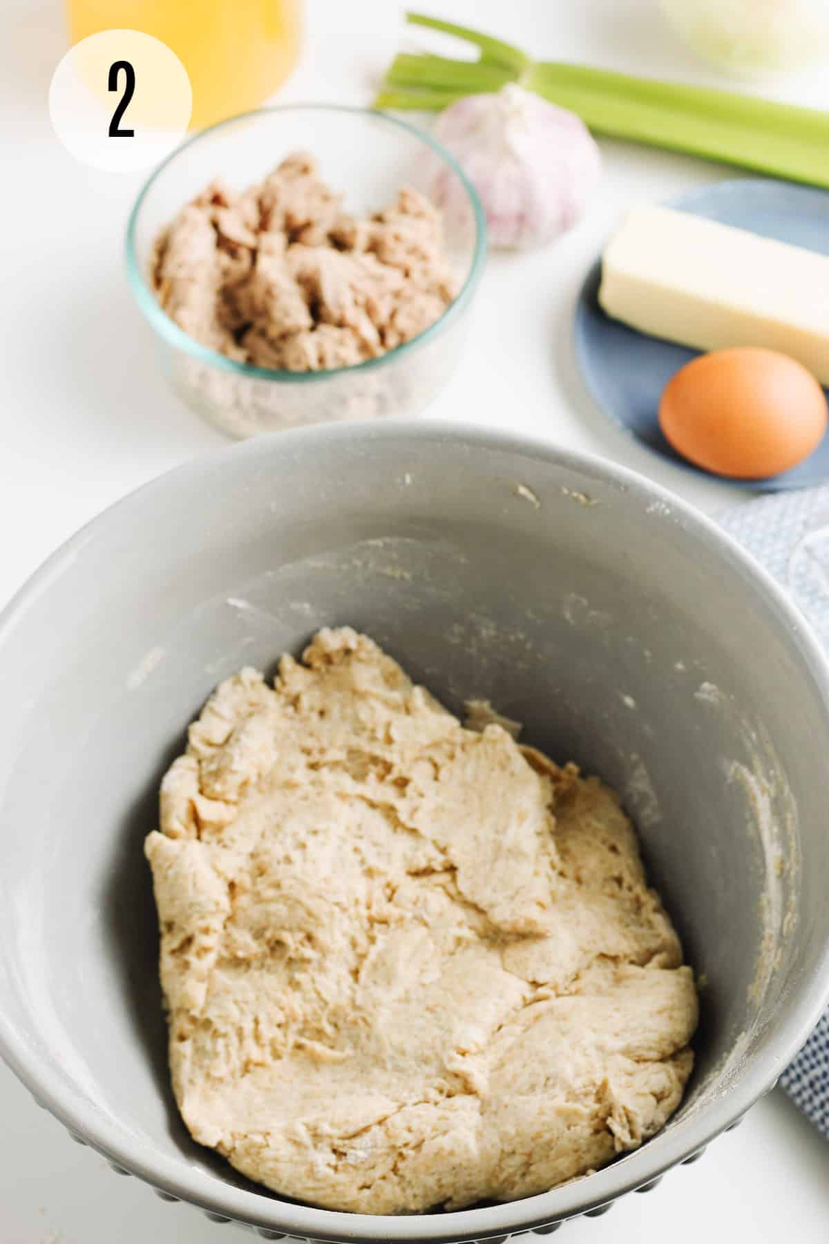 Grey bowl with whole wheat biscuit dough and tuna, garlic, celery stalk, butter and egg in background. 