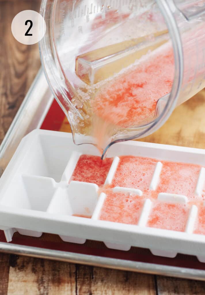 Blender jar pouring pureed watermelon juice into a white ice cube tray for freezing. 