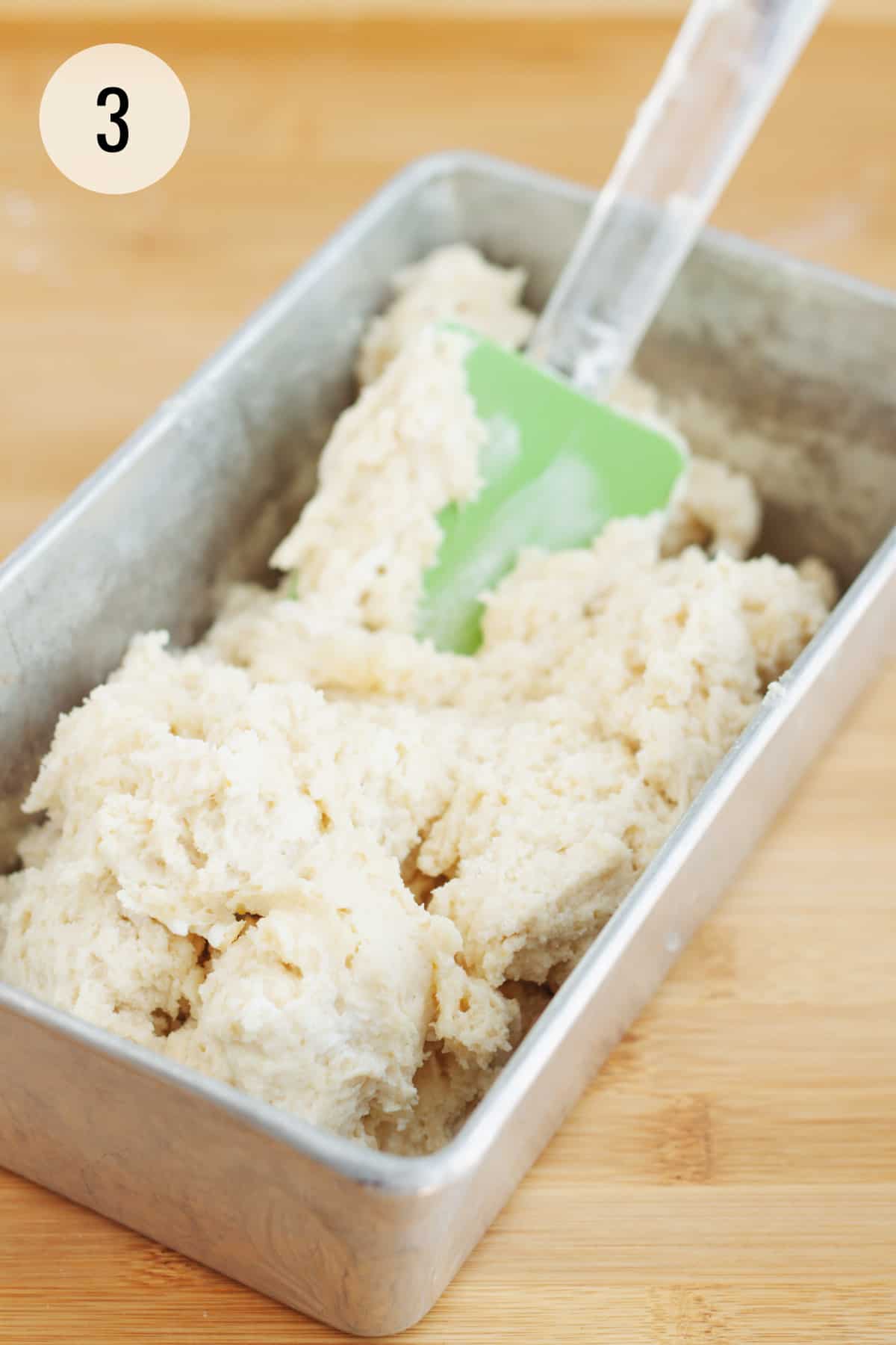 Spreading lemon bread batter in a metal baking pan using a green rubber spatula. 