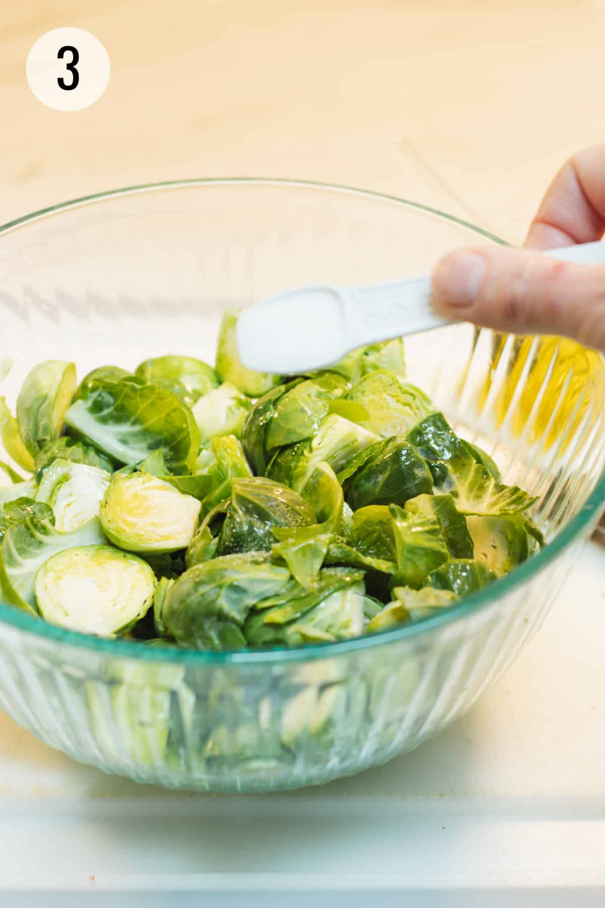 White teaspoon sprinkling salt on glass bowl full of cut Brussels sprouts. 