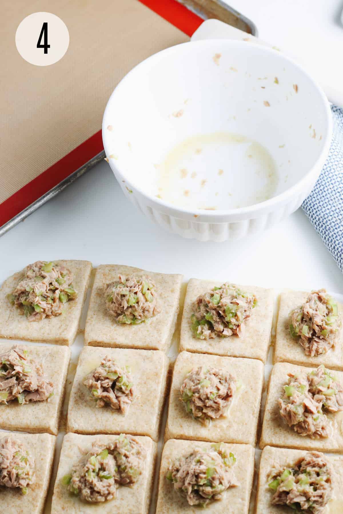 Squares of biscuit dough with scoops of tuna and celery mixture on each and white bowl and baking pan in upper background. 