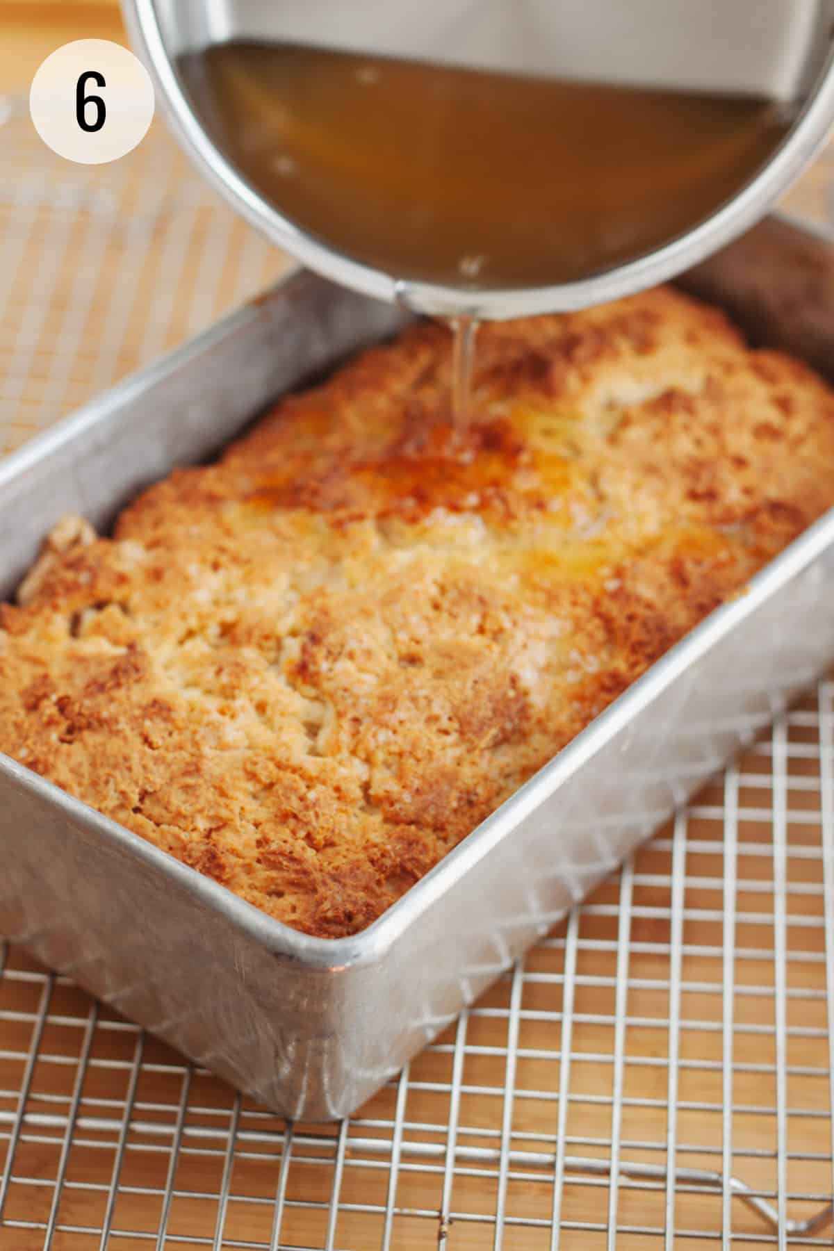 Pouring lemon simple syrup over baked lemon bread in a metal baking pan on cooling rack. 