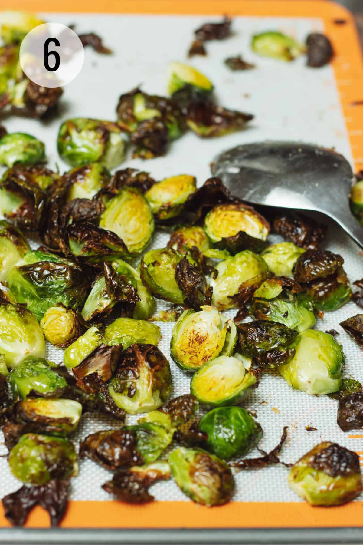 Roasted Brussels sprouts cut in half with crispy brown bits all on a baking sheet with a silver spoon on the right of photo. 