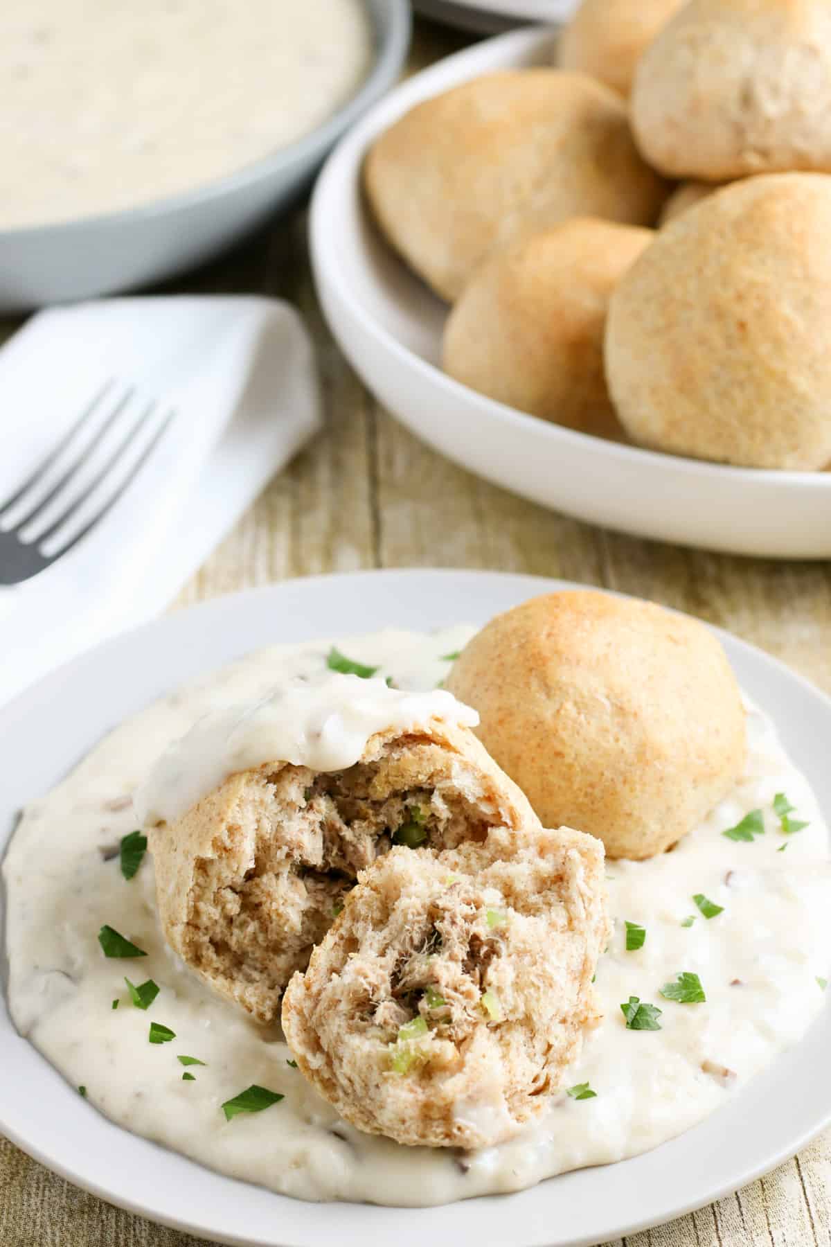 Tuna fish mixture wrapped in a baking powder biscuit on plate withe creamy mushroom gravy sprinkled with green parsley and bowl of pockets and fork in background.
