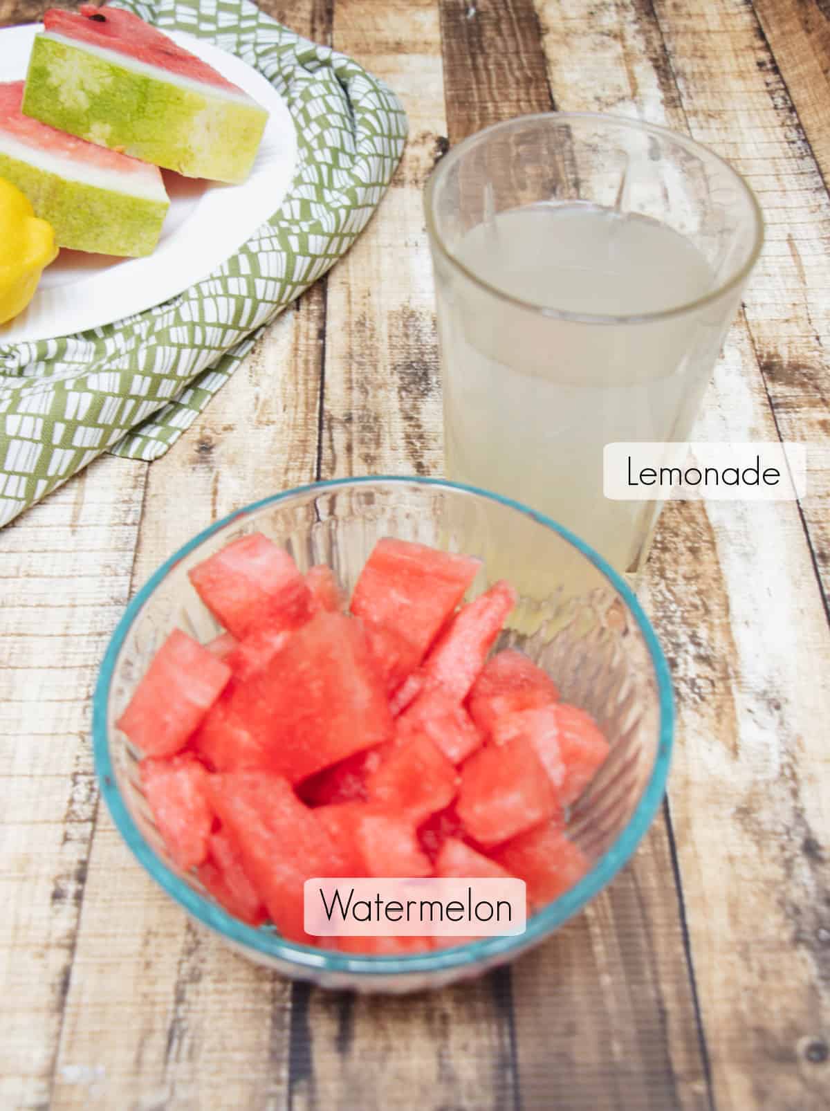 Glass bowl with cubed watermelon and glass of lemonade, both labeled, with plate of watermelon slices and lemon in upper left corner. 