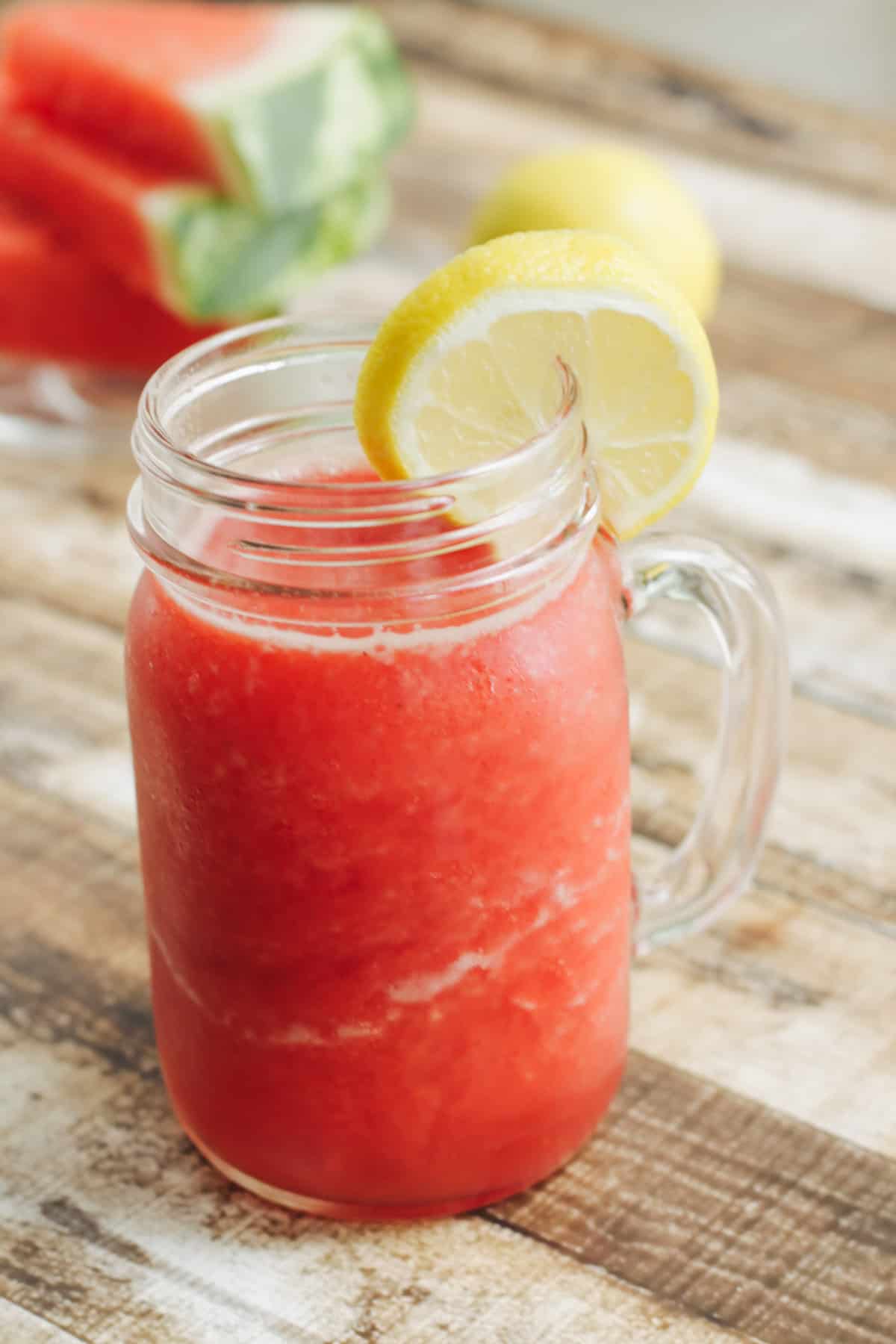 Mason jar style glass mug filled with watermelon lemonade and slice of lemon for garnish and watermelon slices in upper left background.