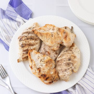 White plate with grilled chicken breasts on a blue and white towel with silver fork to side.