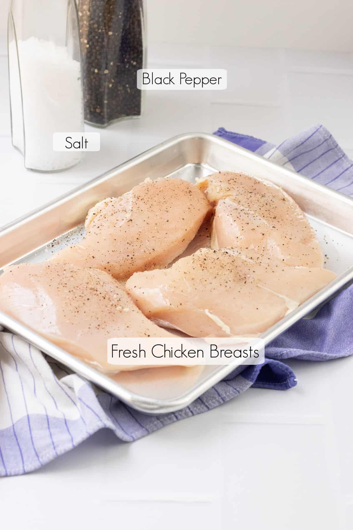 Metal baking tray with uncooked chicken breasts sprinkled with salt and pepper on a blue and white towel with salt and pepper shakers in background. 