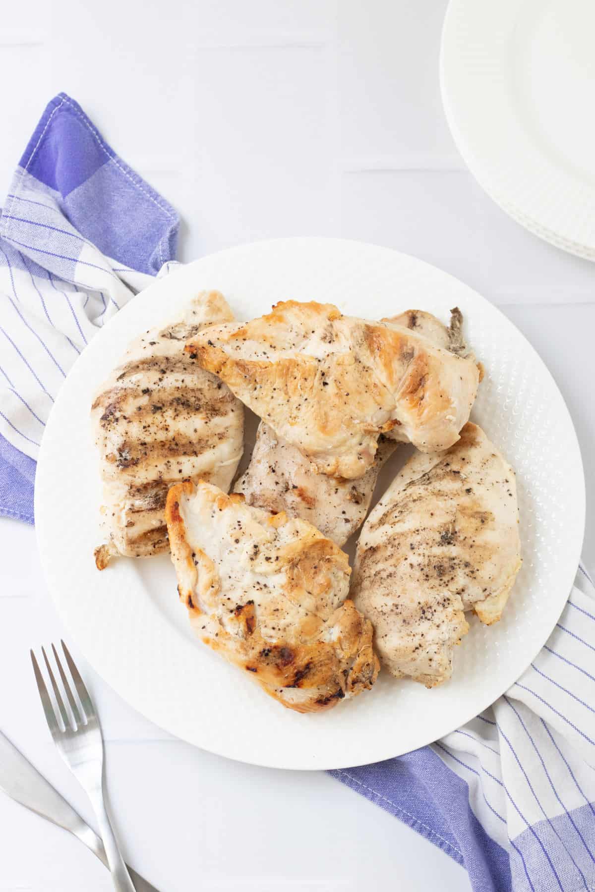 Whole grilled chicken breasts on a white plate on a blue and white towel and fork in lower right corner.