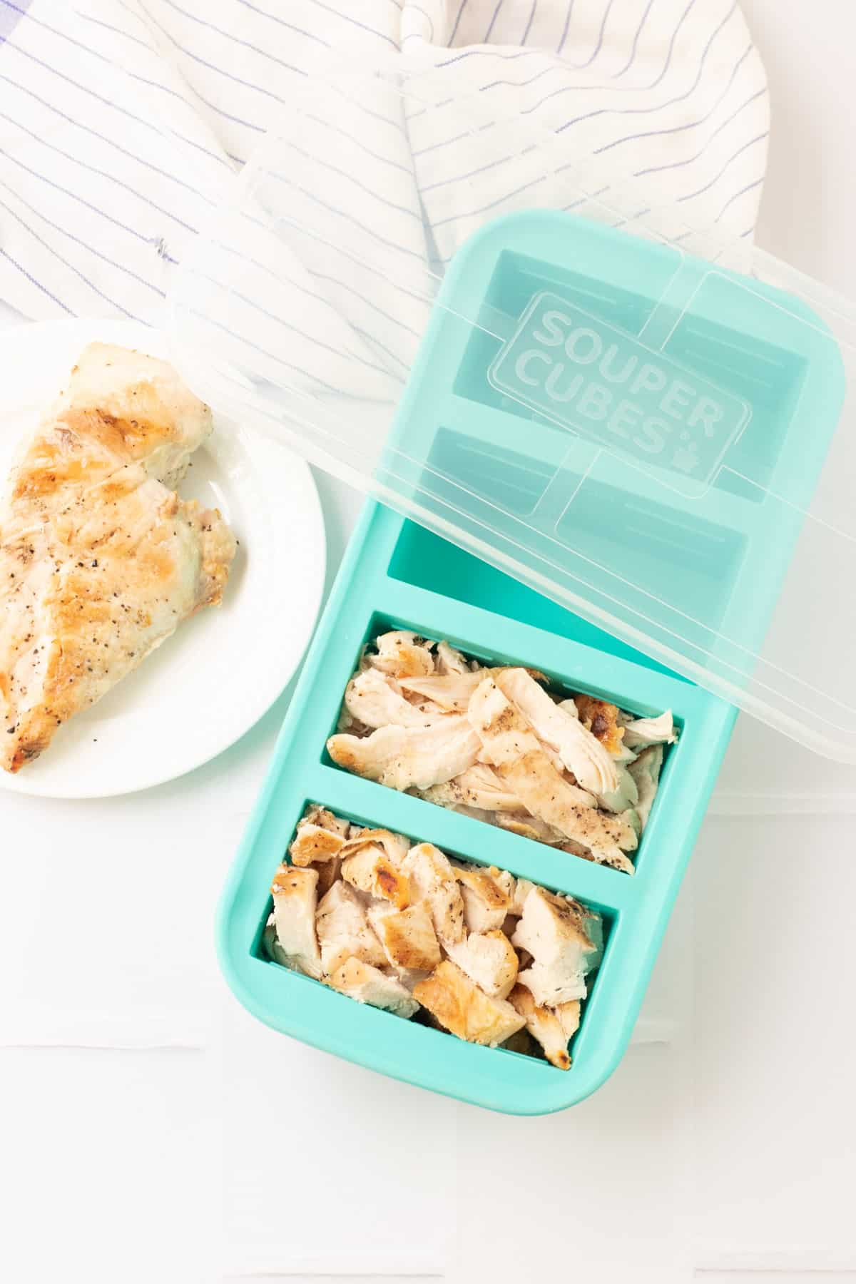 Aqua SouperCubes freezer tray with cubed and shredded chicken and clear lid on upper tray and single chicken breast on plate to left. 