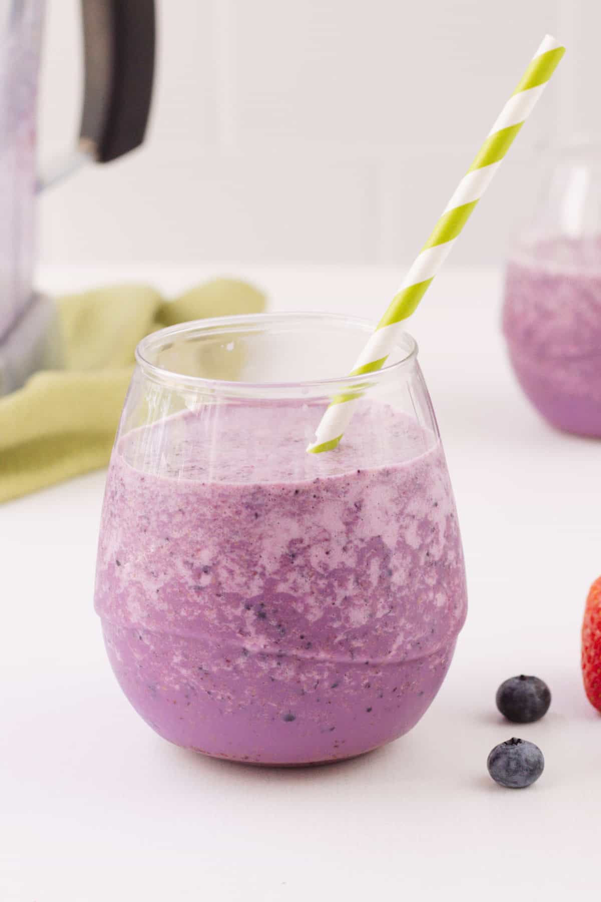 Glass with purple peanut butter and jelly smoothie with green and white striped straw, another glass in right background, blender jar in left background and blueberries in foreground.