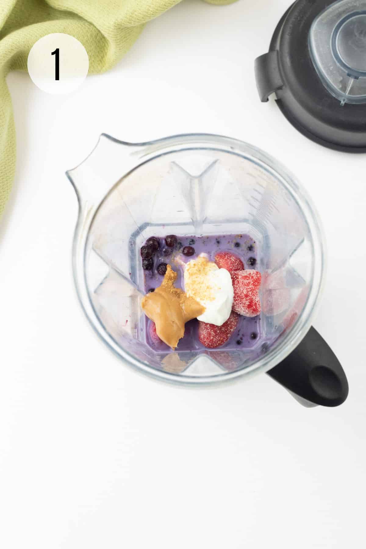 Top view of blender jar with blueberries, strawberries, peanut butter, yogurt and flax seed and blender jar lid in upper right and green linen in upper left.