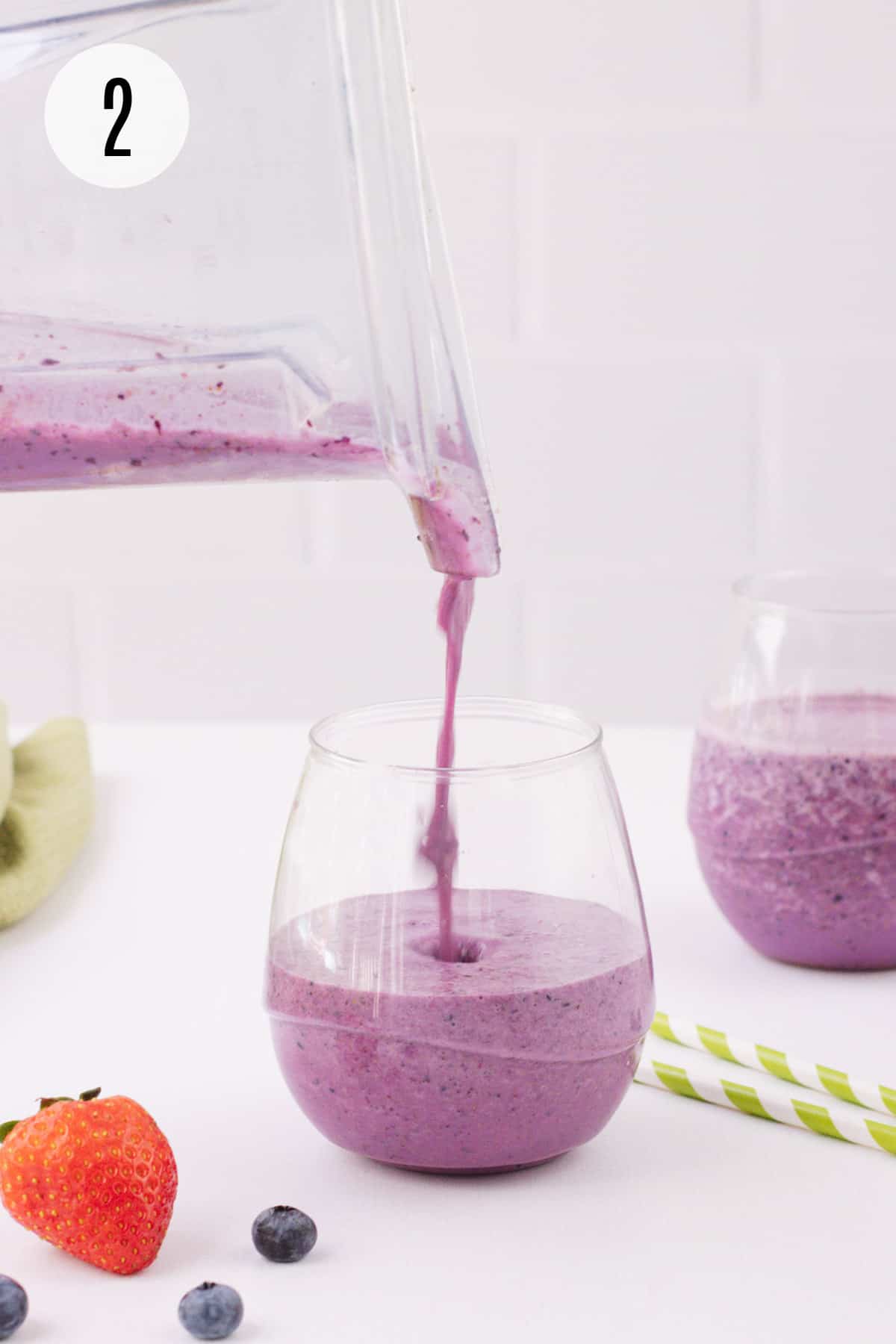 Pouring PB&J Smoothie into glass with strawberry and blueberries in foreground and another glass with smoothie and green and white striped straw in background.