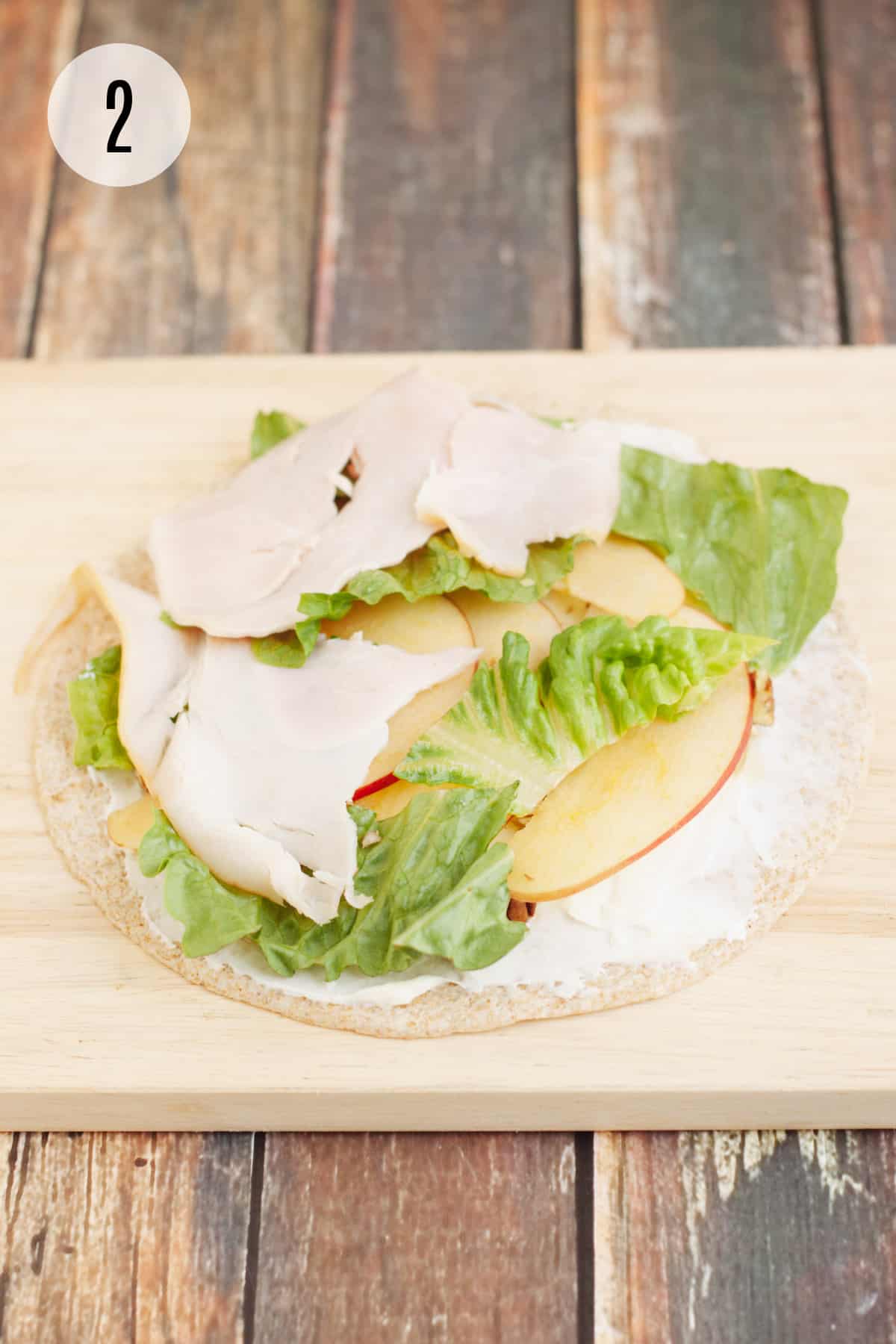 Whole wheat tortilla layered with cream cheese, sliced apples, lettuce and deli turkey on a wooden cutting board and step #2 in upper left corner. 
