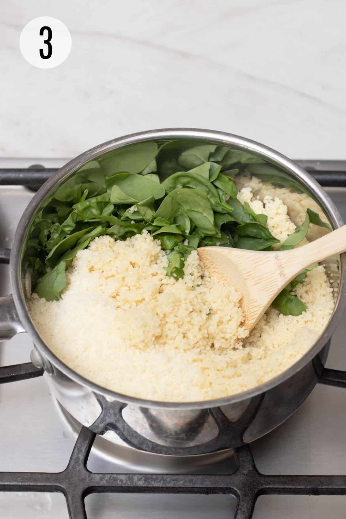 Silver saucepan with wooden spoons stirring chopped baby spinach into couscous. 