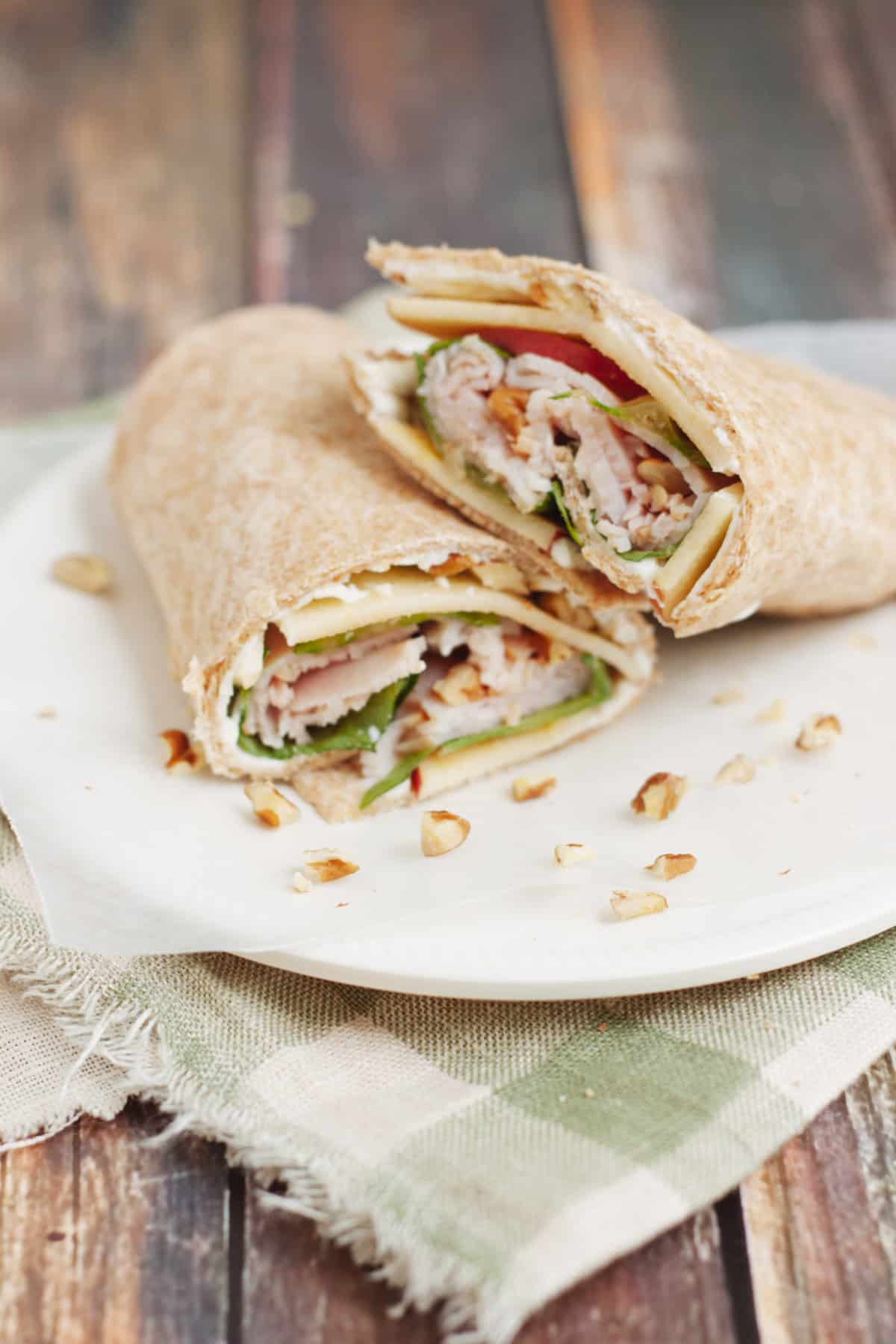 Deli turkey, lettuce and sliced apples wrapped in whole wheat tortilla with pecans scattered on plate on top of green and white check fabric.