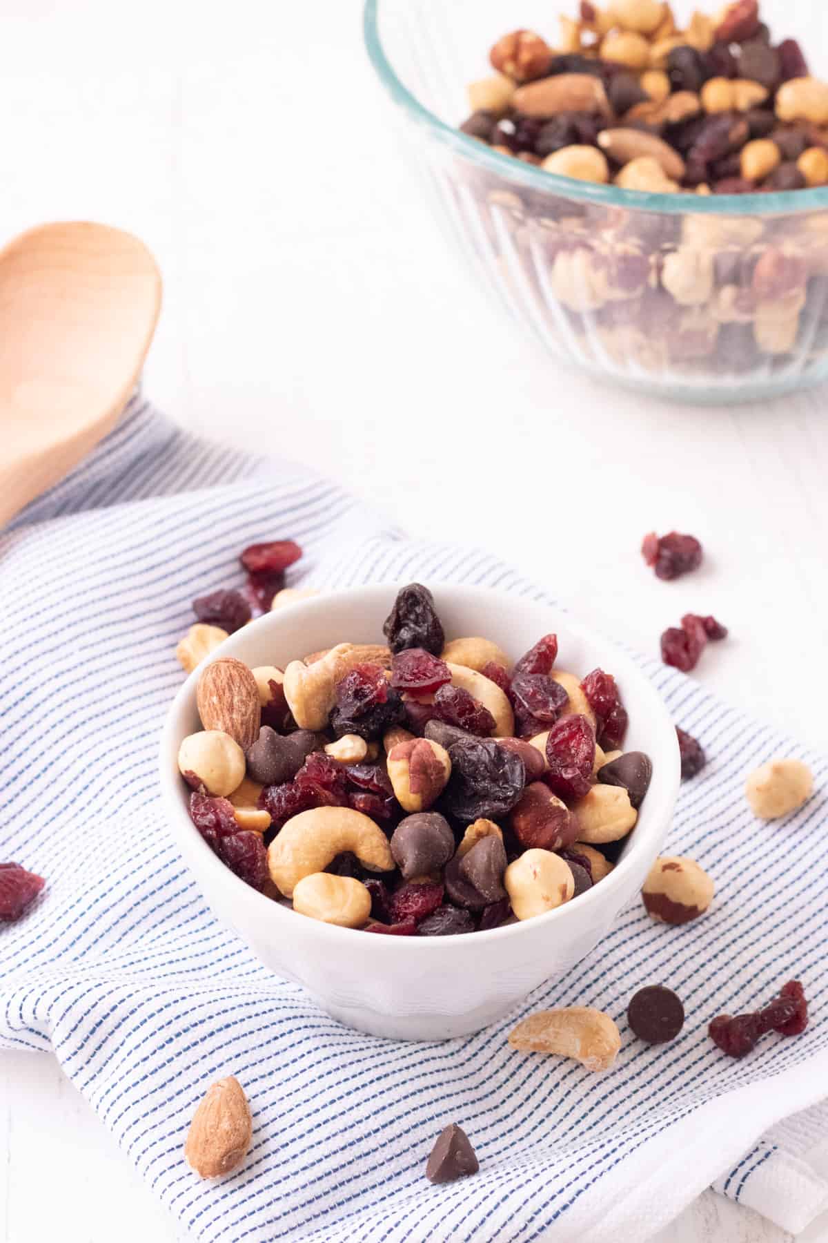 Small white bowl with roasted nuts and dried cherries and cranberries and sprinkled on a blue and white striped towel with wooden spoon and bowl of trail mix in upper background.