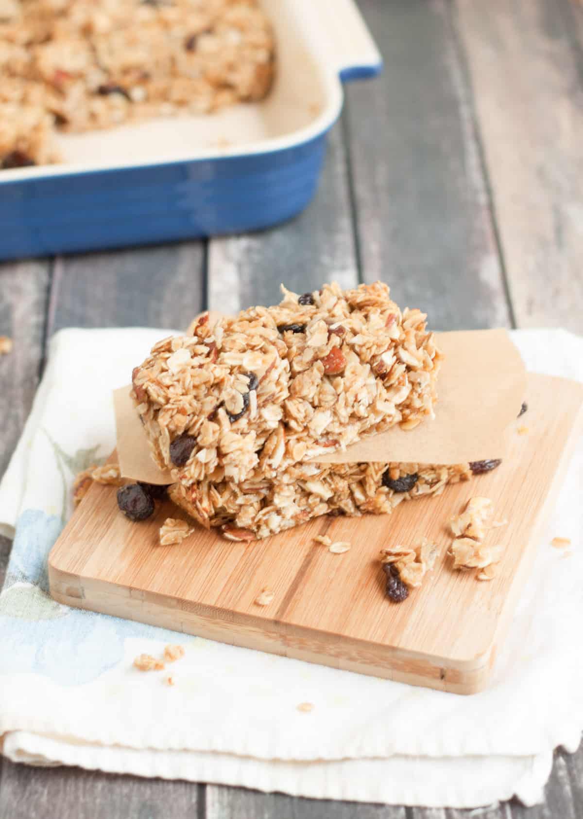 Stack of homemade granola bars on wooden cutting board divided by brown parchment paper with blue baking pan of bars in background. 