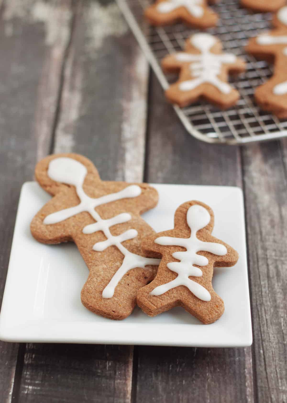 Mexican chocolate sugar cookies decorated to look like skeletons placed on a white plate with extras on a baking rack in upper right. 