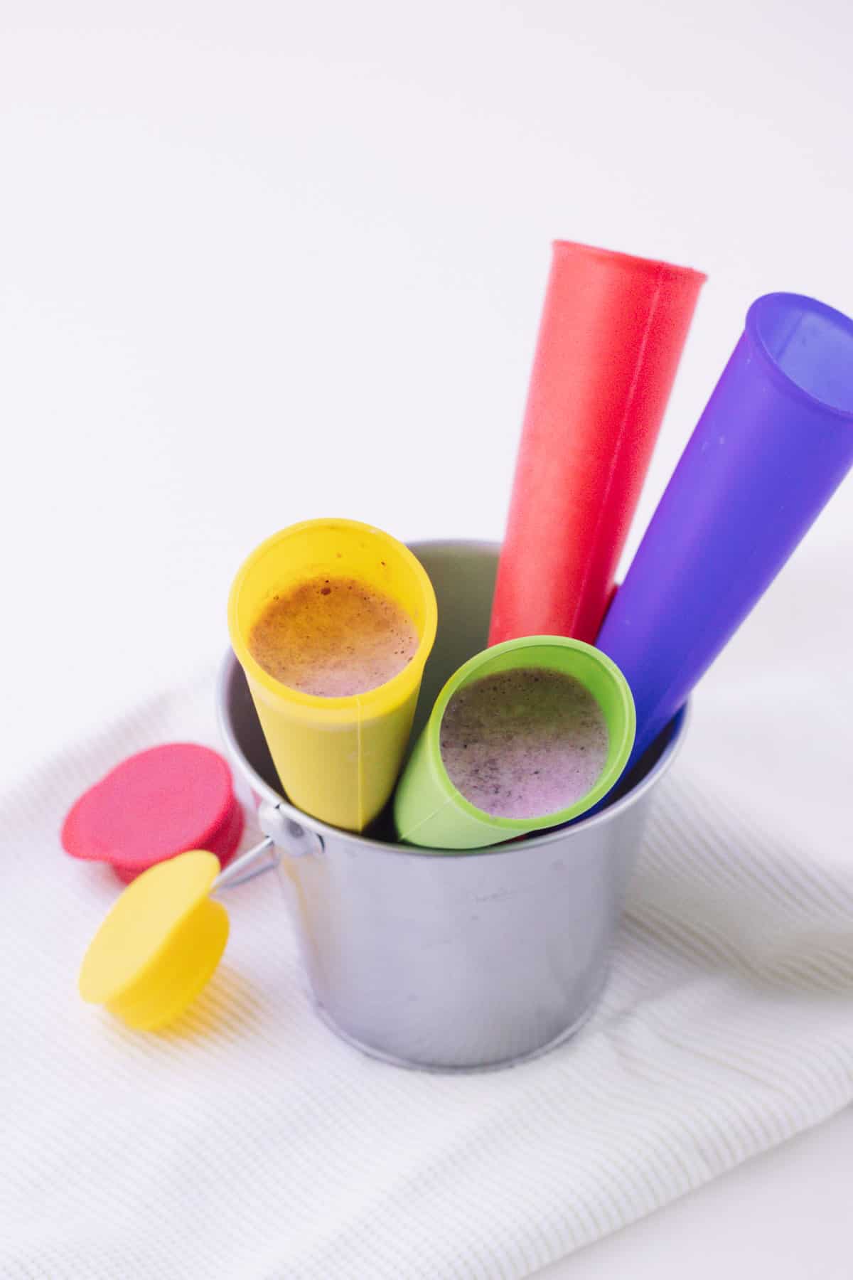 Multi-colored silicone freezer pop molds filled with PB&J smoothie in a small silver bucket and red and yellow lid to left.