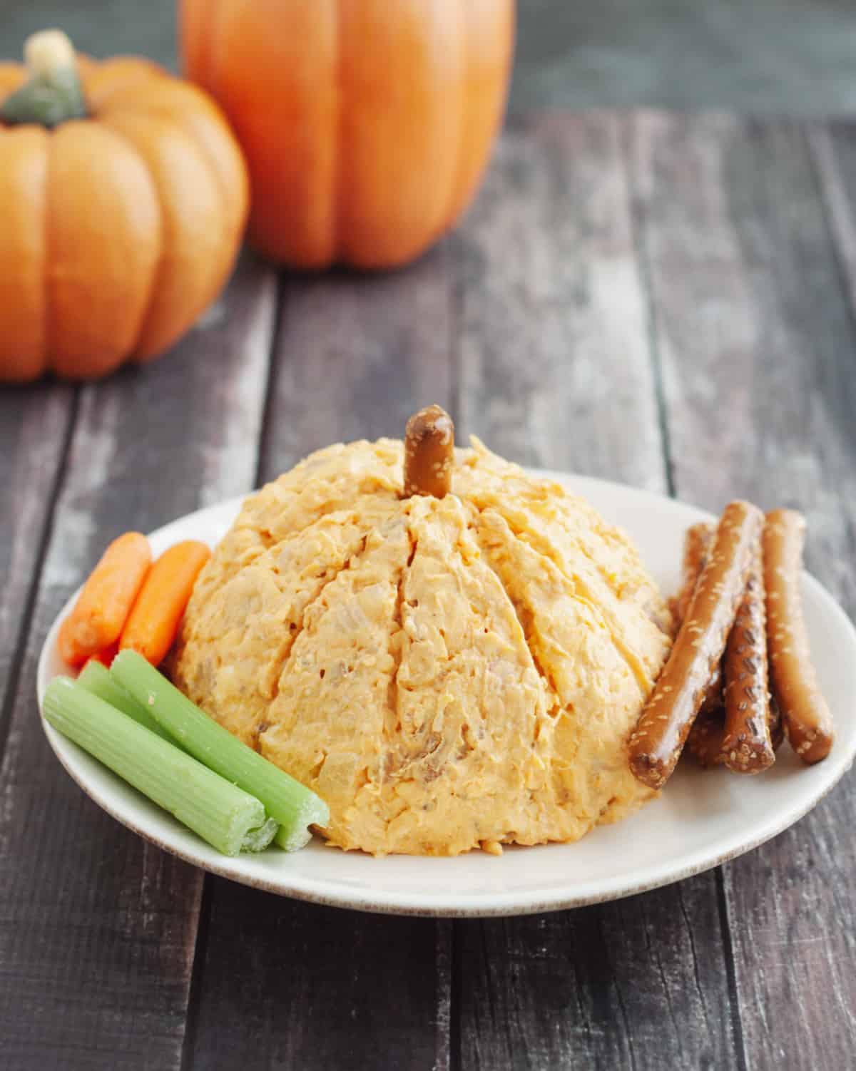 Orange pumpkin shaped cheeseball with celery and carrots and pretzel rods on a white plate with fake pumpkins in upper left background.