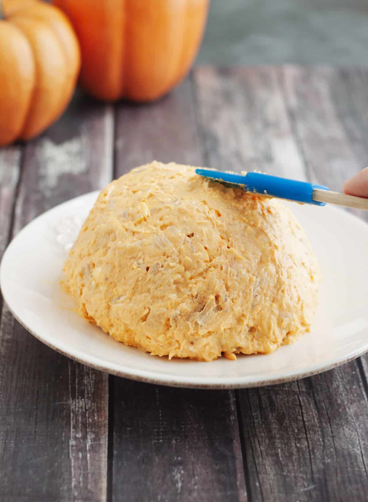 Orange pumpkin cheeseball appetizer spreading with a blue rubber spatula on a white plate with fake pumpkins in upper left background. 