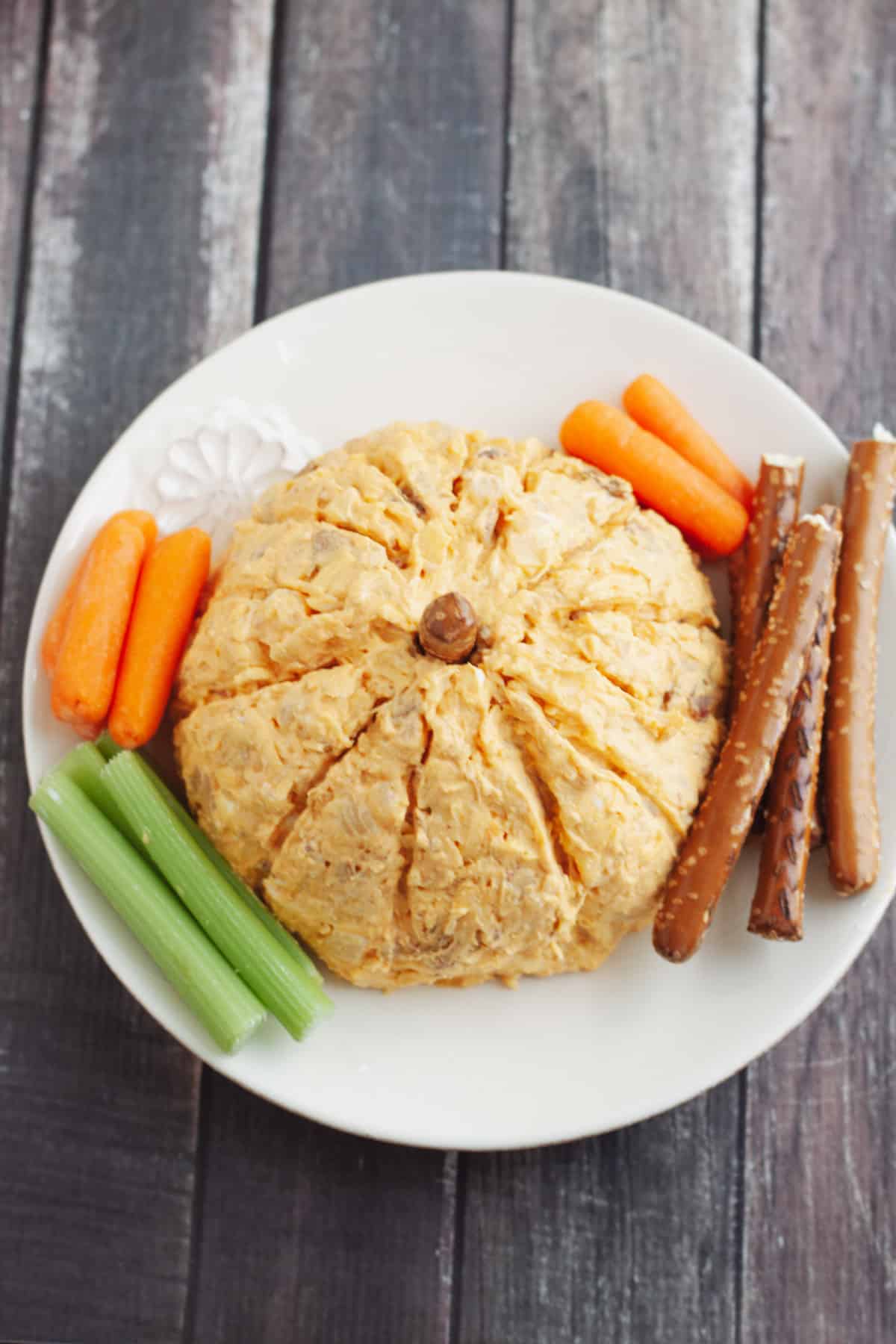 Top view image of pumpkin shaped cheeseball on a white plate with pretzel rod stem and large pretzel rods, celery and carrot sticks for serving. 