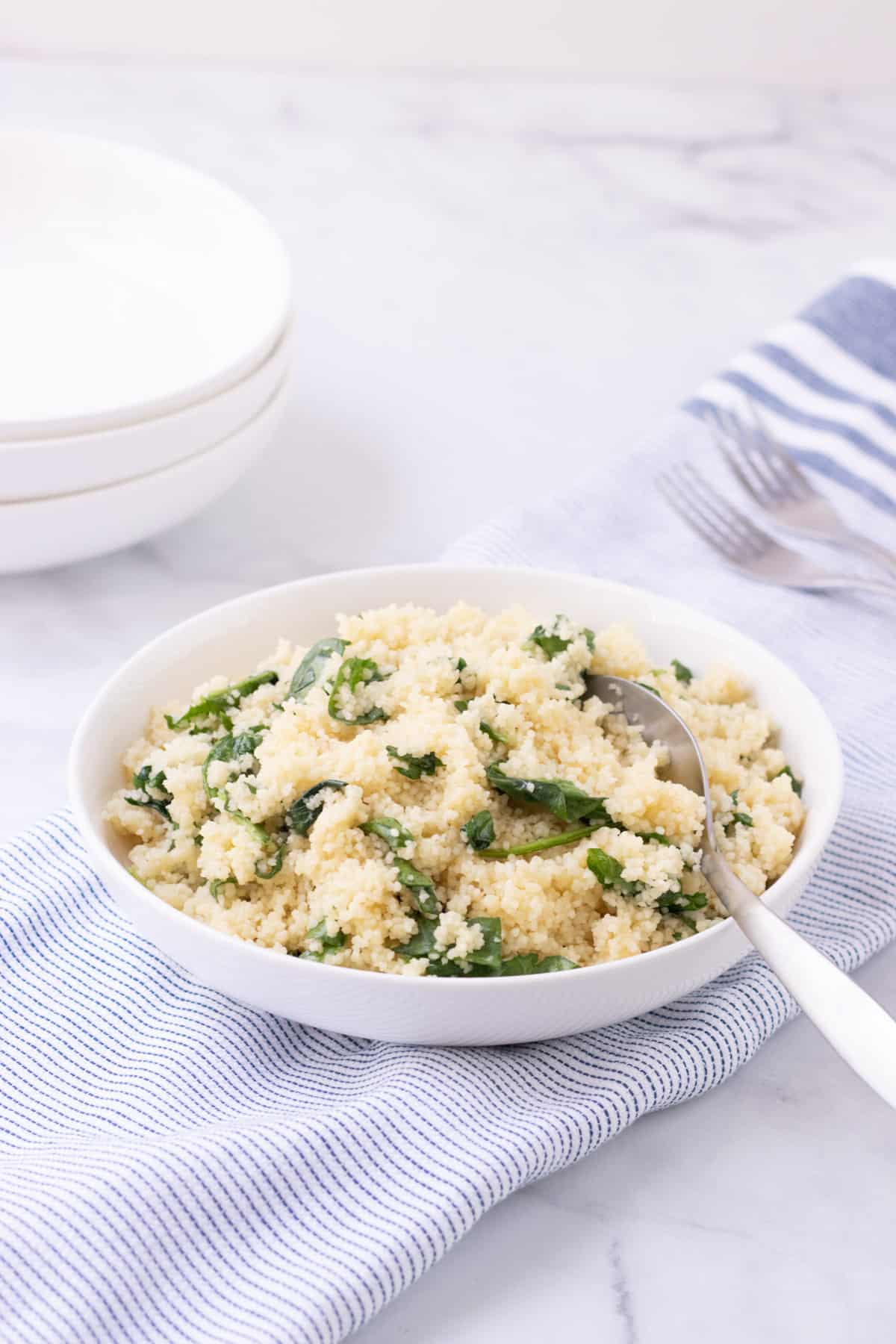 White bowl  with spinach Parmesan couscous on blue and white striped linen with silver spoon and white bowls in left upper background.