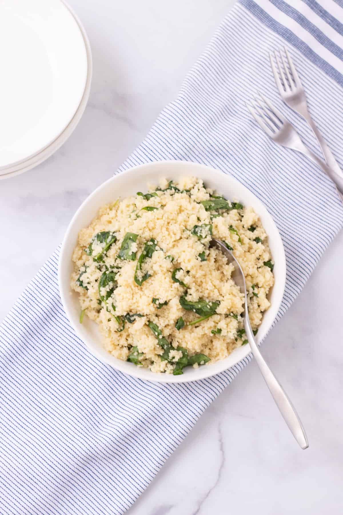 Top view image of spinach parmesan couscous in a white bowl on top of blue and white striped linen with extra bowls and forks in upper background. 