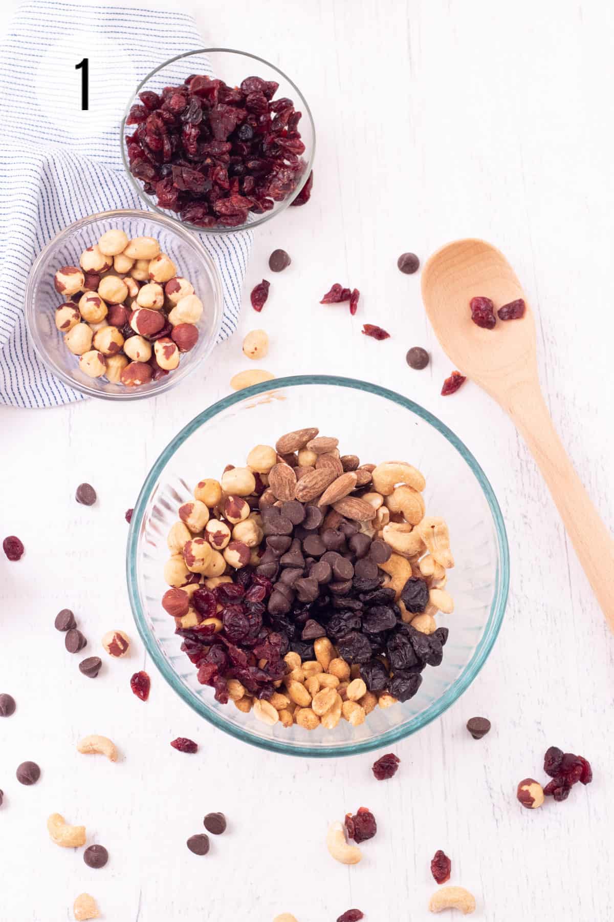 Glass bowl with roasted nuts, dried fruit and chocolate chips and sprinkle of ingredients on background with wooden spoon and smaller bowls of nuts in background. 